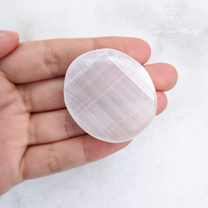 Hand holding a round, white stone with visible layers against a white background. Selenite palm stones available from Inner Nurture, an online crystal store in australia. 